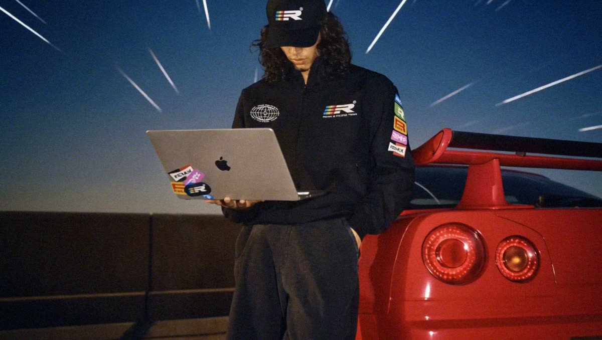 Model wearing a black Remix Racing jacket and hat while holding a laptop beside a red race car